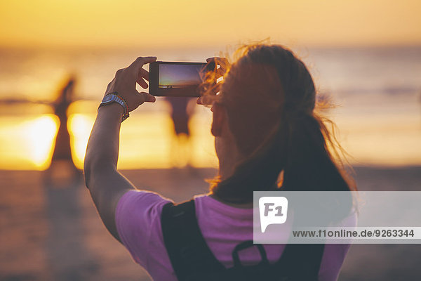 Indonesien  Bali  Frau am Strand fotografiert Sonnenuntergang mit ihrem Smartphone