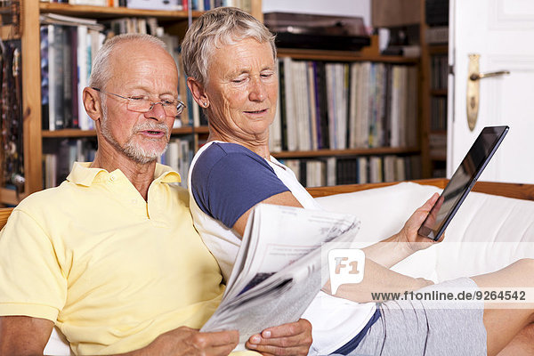 Seniorenpaar sitzt auf der Couch und liest Zeitung mit digitalem Tablett.