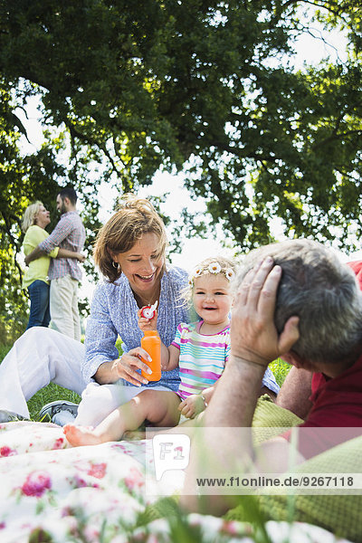 Großeltern mit ihrer kleinen Enkelin auf der Wiese  während sich das junge Paar im Hintergrund umarmt.