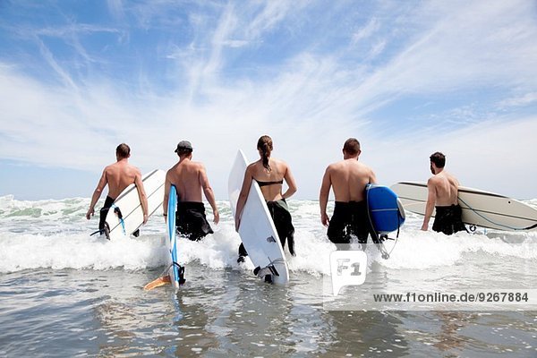 Rückansicht einer Gruppe männlicher und weiblicher Surferfreunde  die mit Surfbrettern ins Meer waten.