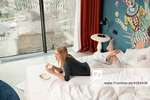Young woman having a coffee whilst lying on hotel bed