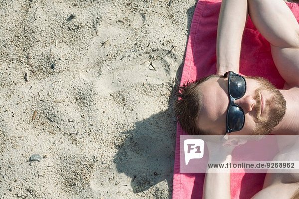 Overhead-Ansicht eines erwachsenen Mannes mit Sonnenbrille beim Sonnenbaden auf Sand