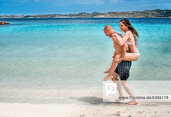 Mid adult man giving girlfriend piggy back on beach  La Maddalena  Sardinia  Italy