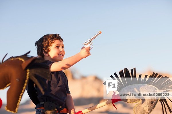 Junge als Cowboy verkleidet mit Hobbypferd und Spielzeugpistole in Sanddünen
