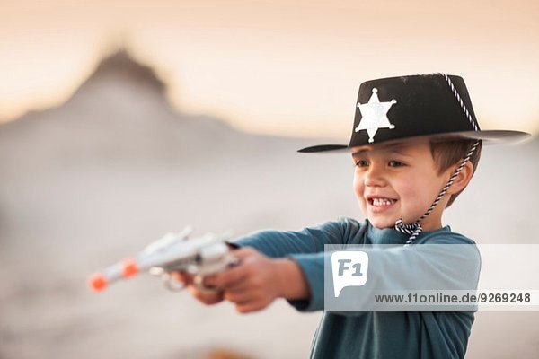 Junge als Cowboy-Sheriff verkleidet  der Spielzeugpistolen in Sanddünen hält.