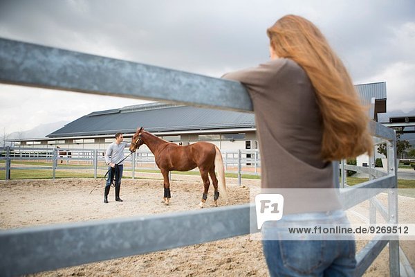 Young woman watching stablehand train horse in paddock ring