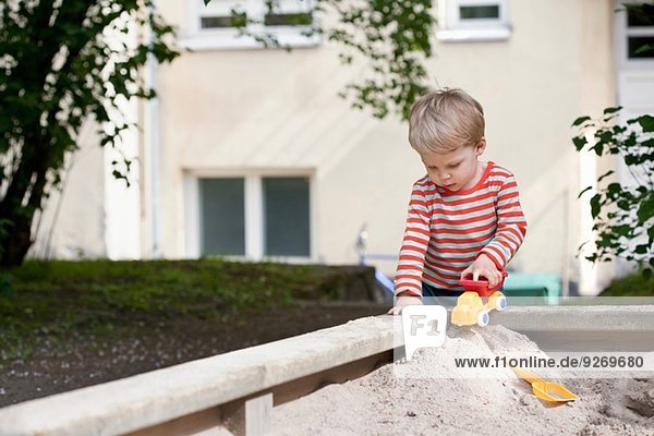 Männliches Kleinkind schiebt Spielzeugauto in Sandkasten im Garten