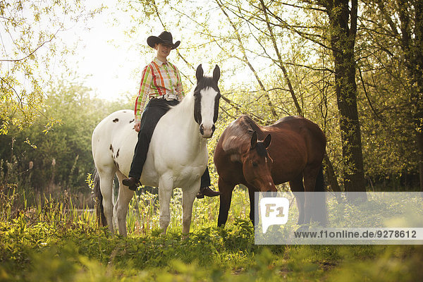 Westernreiterin auf Paint Horse in der Färbung Black Tobiano mit Handpferd brauner Shagya-Araber bei Ausritt ohne Sattel durch Wald  Münsterland  Nordrhein-Westfalen  Deutschland