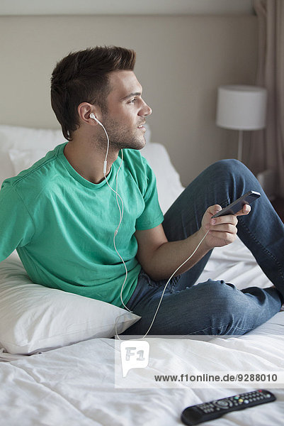 Junger Mann sitzt auf dem Bett und hört Musik mit dem Smartphone.