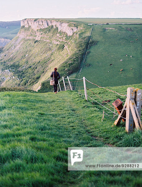 Eine Frau  die entlang einer Zaunlinie auf einem Klippenpfad geht. Blick auf die Landschaft.