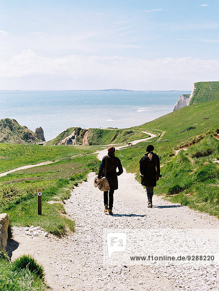 Zwei Frauen gehen auf einem Küstenwanderweg entlang von Klippen.