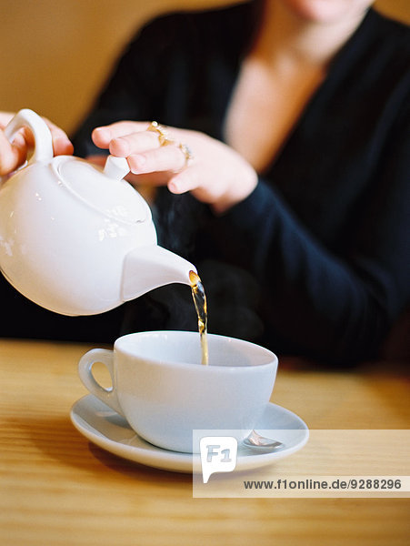 Eine sitzende Frau gießt aus einer Teekanne aus weißem Porzellan eine Tasse Tee aus.