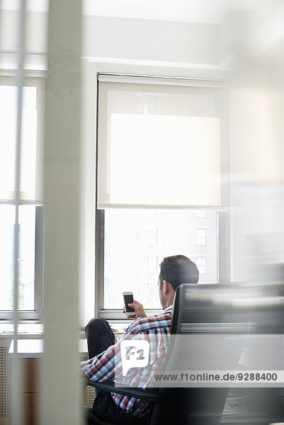 Ein Mann  der in einem großen Bürostuhl sitzt  sich zurücklehnt und sein Smartphone kontrolliert.