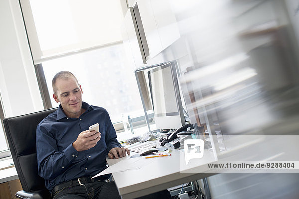 A man sitting at an office desk checking his smart phone.