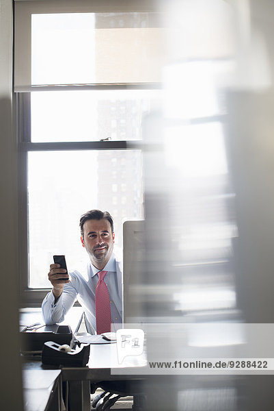 Ein Mann arbeitet an einem Bürocomputer und hält ein Smartphone hoch  um nach Nachrichten zu suchen.