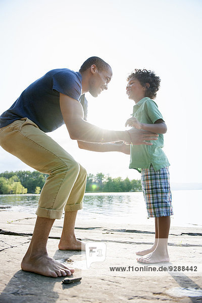 A man lifting a small boy up  playing in the sun by a lake.