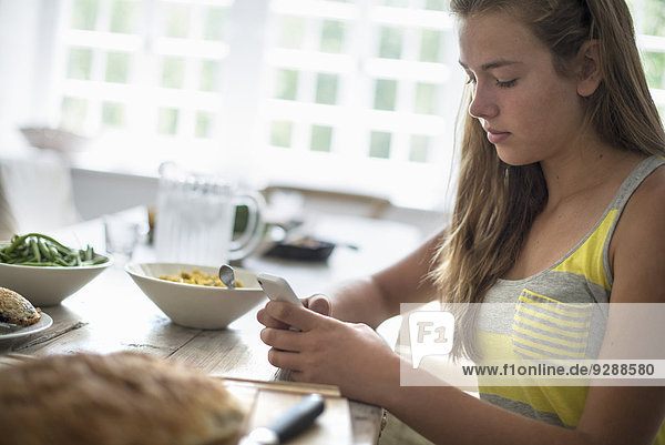A young girl seated checking her smart phone at a dining table.