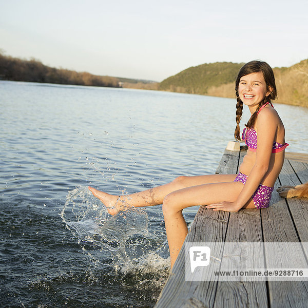 A girl in a bikini sitting on a jetty with her feet in the water.