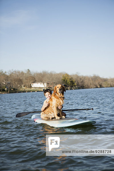 Ein Kind und ein Apportierhund sitzen auf einem Paddelbrett auf dem Wasser.