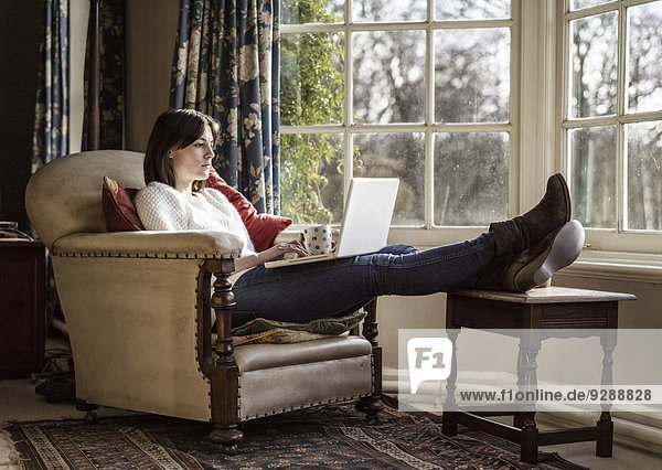 Eine junge Frau  die sich zu Hause mit erhobenen Füßen und einem Laptop entspannt.