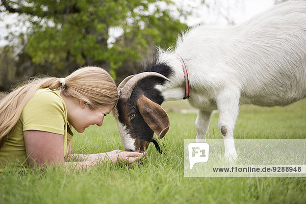 A girl lying on grass head to head with a goat.