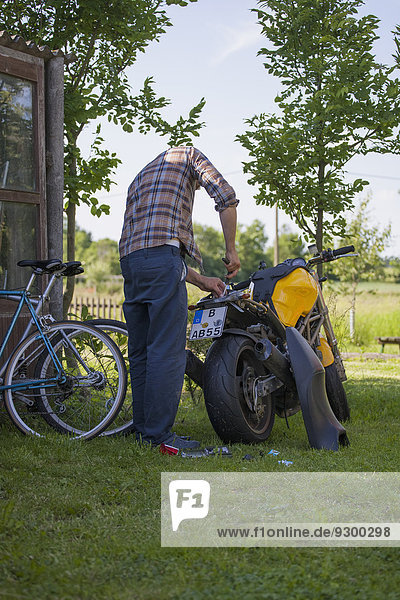 Vollständige Rückansicht des reifen Mannes  der das Motorrad auf dem Hof repariert.