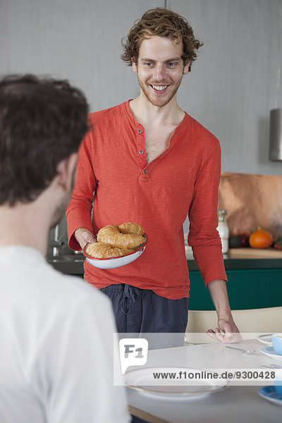 Smiling gay man holding croissants in plate looking at partner at home