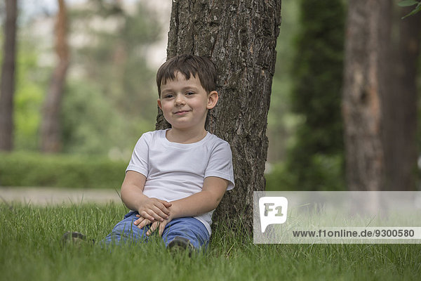 Porträt eines süßen Jungen  der sich gegen einen Baum im Park entspannt.