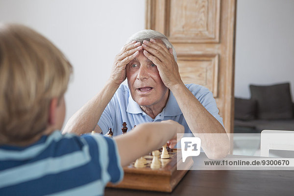 Schockierter älterer Mann mit Kopf in den Händen  der den Enkel beim Schachspielen zu Hause ansieht.
