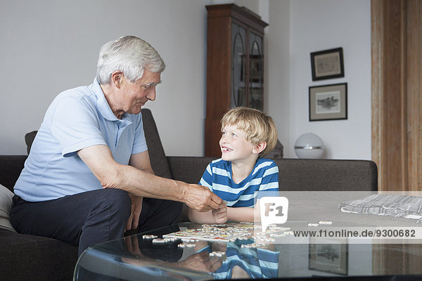 Glücklicher Enkel und Großvater schauen sich an und lösen Puzzle im Wohnzimmer.