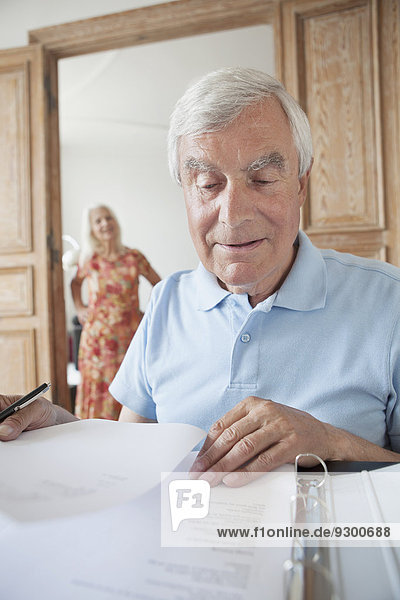 Senior-Mann bei der Prüfung von Finanzdokumenten zu Hause mit Frau im Hintergrund
