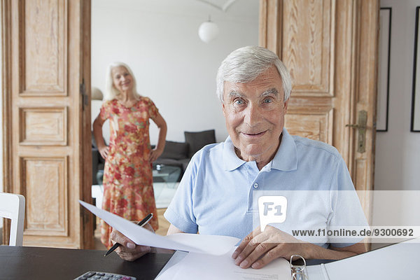 Porträt eines überraschten älteren Mannes  der zu Hause mit einer Frau im Hintergrund Finanzdokumente prüft.