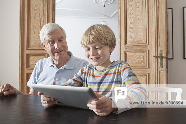 Senior Mann,  der den Enkel mit einem digitalen Tablett im Haus betrachtet.