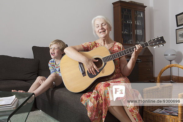 Playful boy sitting with grandmother singing while playing guitar on sofa at home