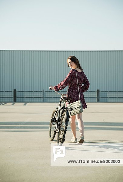 Junge Frau schaut zurück  während sie das Fahrrad auf dem leeren Parkplatz schiebt.