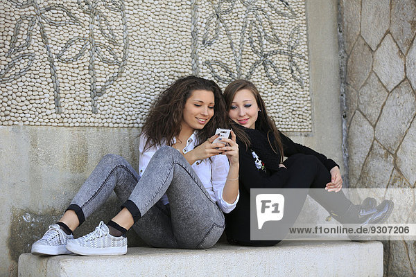 Zwei Freundinnen  Teenager  sitzen Rücken an Rücken auf einer steinernen Bank und lesen eine SMS auf einem Handy  Menton  Provence-Alpes-Côte d?Azur  Frankreich