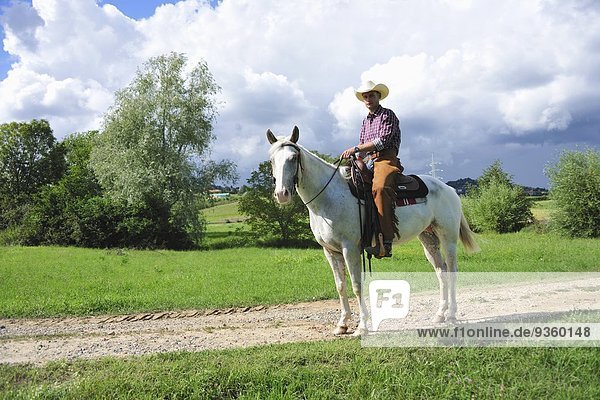 Porträt eines jungen Mannes in Cowboy-Ausrüstung zu Pferd auf Feldweg