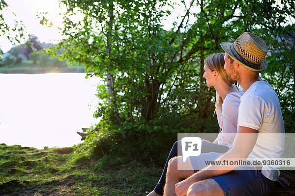 Mittleres erwachsenes Paar  das am See sitzt und den Sonnenuntergang beobachtet.
