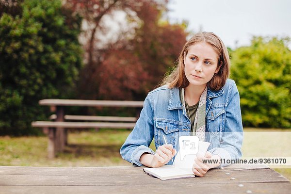 Junge Frau sitzt auf einer Picknickbank im Park und schreibt in ein Notizbuch.