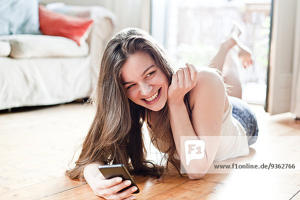 Porträt einer lächelnden jungen Frau mit Smartphone im Wohnzimmer