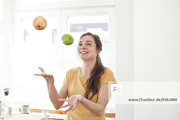 Lächelnde junge Frau jongliert mit zwei Äpfeln in ihrer Küche