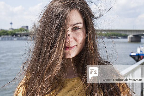 Deutschland  Köln  Porträt einer lächelnden jungen Frau mit wehendem Haar vor dem Rhein stehend
