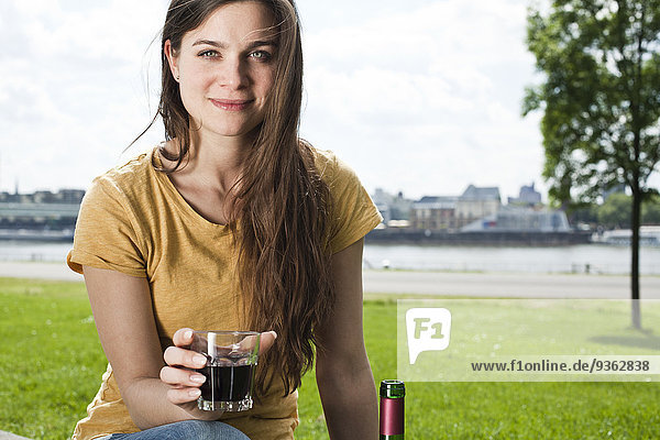 Deutschland  Köln  Porträt einer lächelnden jungen Frau mit einem Glas Rotwein vor dem Rhein