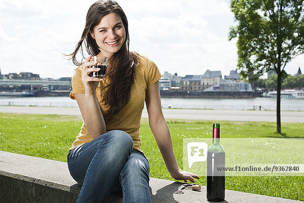Deutschland  Köln  Porträt einer lächelnden jungen Frau  die vor dem Rhein sitzt und Rotwein trinkt.