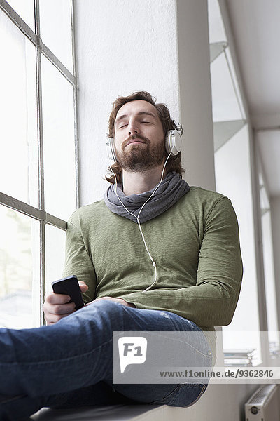 Portrait of young man sitting on window sill in an office listening music with headphones