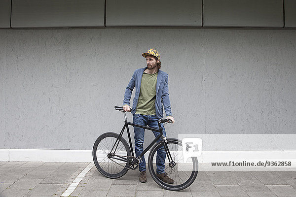 Germany  Bavaria  Munich  young man wearing basecap standing in front of a wall with his racing cycle
