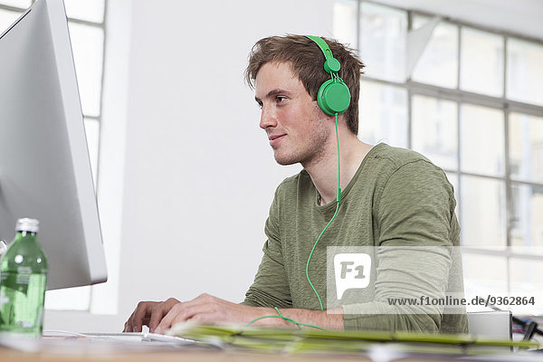 Porträt eines lächelnden jungen Mannes  der am Computer arbeitet  während er mit Kopfhörern Musik hört.