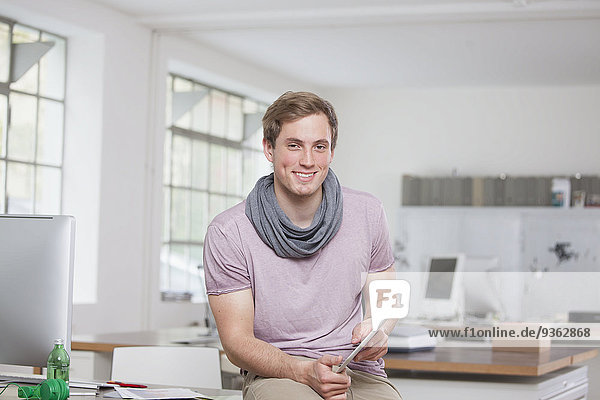 Portrait of smiling young man sitting on his desk in the office holding digital tablet