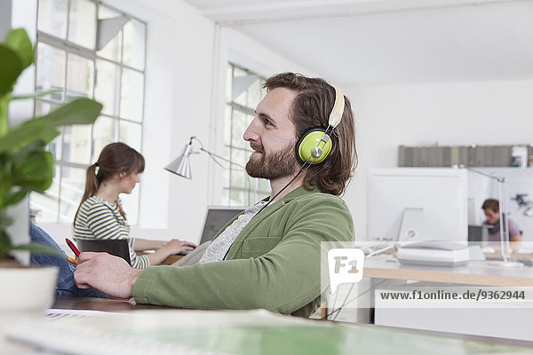 Junger Mann sitzt an seinem Schreibtisch in einem kreativen Büro und hört Musik mit Kopfhörern.