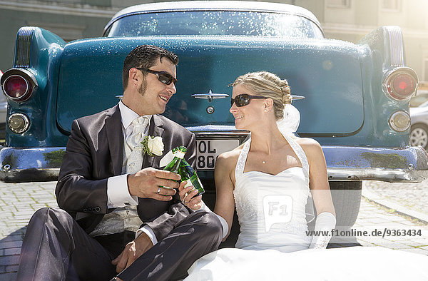 Bridal couple sitting in front of vintage car toasting with piccolo